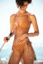 Beautiful girl in bikini using hose pipe on beach. Portrait of lady in beige swimsuit rinsing beach sand off her body on