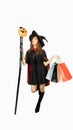 Beautiful girl in black dress and witch hat is holding shopping bags, looking a pumpkin and holding staff on background white. Royalty Free Stock Photo