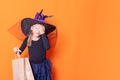 A beautiful girl in a black dress and a witch hat holds a craft paper bag, covering her face with her hand, wondering Royalty Free Stock Photo