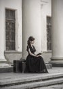 Beautiful girl in black evening dress sitting reading a book. A girl sits with suitcases near an old building with columns. Royalty Free Stock Photo