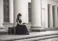 Beautiful girl in black evening dress sitting reading a book. A girl sits with suitcases near an old building with columns. Royalty Free Stock Photo