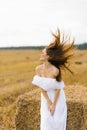 A beautiful girl with blond hair in a white dress sits by a stack of straw in the field. Wind in the hair