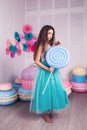 Beautiful girl in blue dress hold in hand huge candy in studio with decor of macaroons. Copy space.