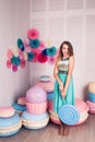 Beautiful girl in blue dress hold in hand huge candy in studio with decor of macaroons. Copy space.