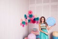 Beautiful girl in blue dress hold in hand huge candy in studio with decor of macaroons. Copy space.