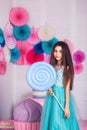 Beautiful girl in blue dress hold in hand huge candy in studio with decor of macaroons. Copy space.