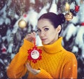 A beautiful girl with blue eyes in a yellow sweater is holding a New Year`s toy under a snow-covered tree. A girl in
