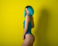Beautiful girl in a blue wig and a bright bikini posing on a yellow background. Woman with artificial hair and a