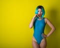 Beautiful girl in a blue wig and a bright bikini posing on a yellow background. Woman with artificial hair and a