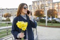 Beautiful girl with bouquet of yellow spring flowers, young woman talking on the phone, urban style background Royalty Free Stock Photo