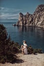 A beautiful girl, a bride, in a white dress sits a tree, on a cliff against the background of the ocean and mountains.