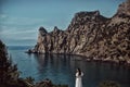 A beautiful girl, a bride, in a white dress stands on a cliff against the background of the sea and mountains.