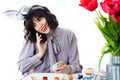 Beautiful girl in bunny ears talking on phone and decorating easter egg at table with paint, brushes, tulips in vase. Stylish Royalty Free Stock Photo