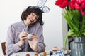 Beautiful girl in bunny ears talking on phone and smiling, painting easter egg at table with tulips in vase. Stylish young Royalty Free Stock Photo