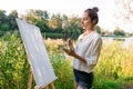 Beautiful girl close-up, woman artist, summer by lake, river pond, draws picture, palette with paints, paintbrush white