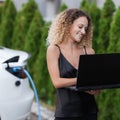 Beautiful girl with curly hair in black outfit working on laptop. Business woman with electric car charging in the background Royalty Free Stock Photo