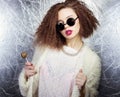 Beautiful girl with curly hair and bright lips in a white coat in the round sunglasses with a candy in his hands, Studio shot