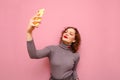 Beautiful girl with curly red hair stands on a pink background and takes a selfie on the camera of a smartphone. Attractive lady Royalty Free Stock Photo