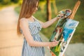 Beautiful girl draws a picture in the park using a palette with paints and a spatula. Easel and canvas with a picture. Summer is a Royalty Free Stock Photo