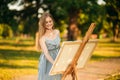 Beautiful girl draws a picture in the park using a palette with paints and a spatula. Easel and canvas with a picture. Summer is a Royalty Free Stock Photo