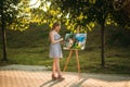 Beautiful girl draws a picture in the park using a palette with paints and a spatula. Easel and canvas with a picture. Summer is a Royalty Free Stock Photo