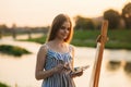 Beautiful girl draws a picture in the park using a palette with paints and a spatula. Easel and canvas with a picture. Summer is a Royalty Free Stock Photo