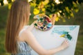 Beautiful girl draws a picture in the park using a palette with paints and a spatula. Easel and canvas with a picture. Summer is a