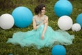 Beautiful girl graduate in a blue dress is sitting on the grass near a large blue and white balloons. Cheerful elegant Royalty Free Stock Photo