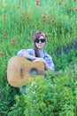 Beautiful girl with a guitar in poppies. Young woman in sunglasses on the grass. A teenager in a denim jacket and a red Royalty Free Stock Photo