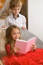 Girl with her hair in red dress is reading pink book sitting in wicker chair. Her brother looks over her shoulder Royalty Free Stock Photo