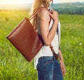 Beautiful girl holding brown leather hand bag outdoors on the sunny meadow at sunset time. Girl wearing fashionable white t-shirt