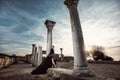 A beautiful girl in a long black dress poses at sunset amid the sea, antique columns and ruins. Royalty Free Stock Photo