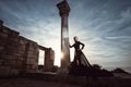 A beautiful girl in a long black dress poses at sunset amid the sea, antique columns and ruins. Royalty Free Stock Photo