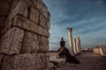 A beautiful girl in a long black dress poses at sunset amid the sea, antique columns and ruins. Royalty Free Stock Photo