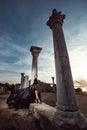 A beautiful girl in a long black dress poses at sunset amid the sea, antique columns and ruins. Royalty Free Stock Photo