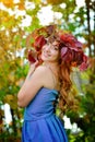 Beautiful girl with long hair in blue dress, in a wreath of red and green leaves standing against bright fall bushes Royalty Free Stock Photo