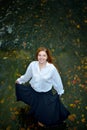 Beautiful girl with long hair in white shirt and black skirt dancing in the river with autumn leaves, looking up and smiling. Royalty Free Stock Photo