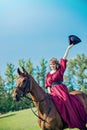 A beautiful girl in a long red dress rides a horse and wags her hat.