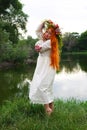 Beautiful girl with long red hair and blue eyes with wreath flowers in white and red embroidered ethnic suit stands by river Royalty Free Stock Photo