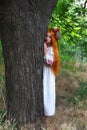 Beautiful girl with long red hair with flower crown in white and red embroidered ethnic suit stands by tree Royalty Free Stock Photo