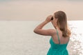 Girl looking in binoculars at sea on twilight. Young woman in turquoise dress observes seascape in cloud weather. Hope and sadness