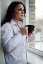 Beautiful girl looks out of window and holds a cup. Winter morning.