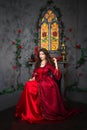 A beautiful girl in a magnificent red dress of the Rococo era sits on a chair near the fireplace, window and flowers.