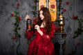 A beautiful girl in a magnificent red dress of the Rococo era stands against a fireplace, a window and flowers with a lamp with ca