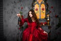 A beautiful girl in a magnificent red dress of the Rococo era stands against a fireplace, a window and flowers with a lamp with ca Royalty Free Stock Photo