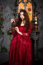 A beautiful girl in a magnificent red dress of the Rococo era stands against a fireplace, a window and flowers with a lamp with ca Royalty Free Stock Photo