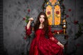 A beautiful girl in a magnificent red dress of the Rococo era stands against a fireplace, a window and flowers with a lamp with ca