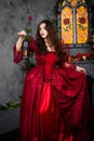 A beautiful girl in a magnificent red dress of the Rococo era stands against a fireplace, a window and flowers with a lamp with ca Royalty Free Stock Photo