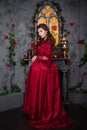 Beautiful girl in a magnificent, red rococo dress. Against the background of the fireplace, window, and flowers.