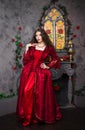 Beautiful girl in a magnificent, red rococo dress. Against the background of the fireplace, window, and flowers. Royalty Free Stock Photo